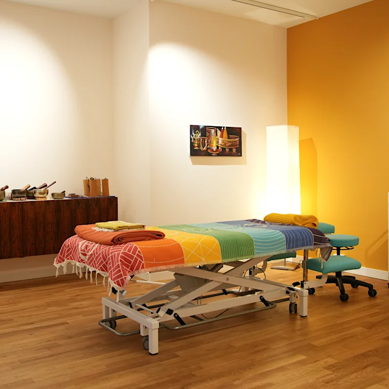 Massageraum, gedämpftes Licht, gelbe Wand, elektrische Massageliege und Klangschalen
